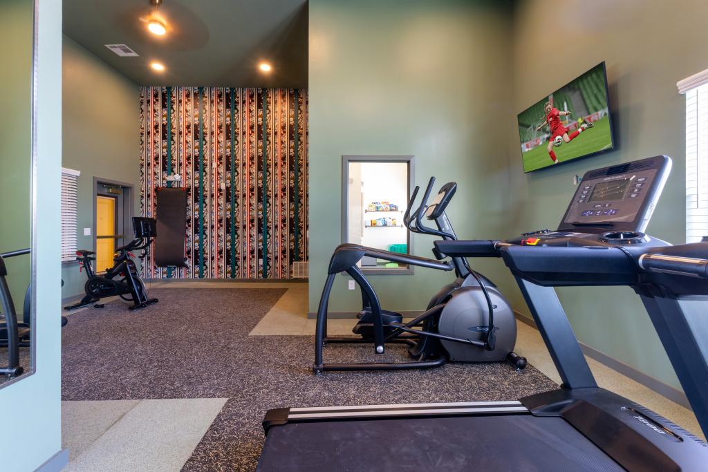A small fitness room with exercise equipment, including a treadmill, an elliptical, and a stationary bike. The walls are painted green, with one accent wall featuring a floral and striped pattern. A TV mounted on the wall displays a soccer game. 