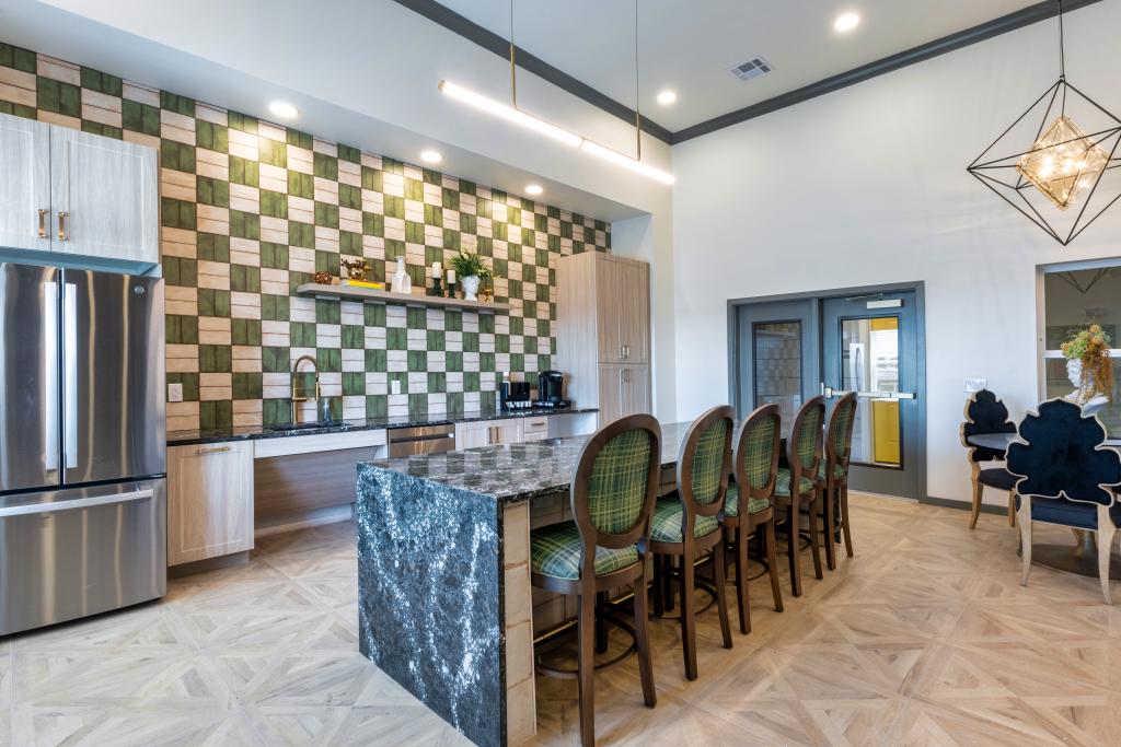 A spacious, modern community kitchen with a long island featuring a dark marble countertop and high-backed green upholstered chairs. The backsplash has a checkered pattern with shades of green and beige. The kitchen includes stainless steel appliances, light wooden cabinets, and a minimalist pendant light above the island. 