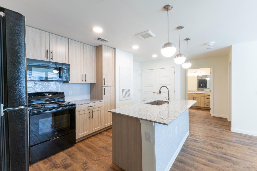 A contemporary apartment kitchen with light wood cabinets, a granite countertop, and a black appliance set, including a stove and refrigerator. Pendant lights hang over the island, and there is a hexagonal backsplash in soft gray tones, giving the space a sleek look.