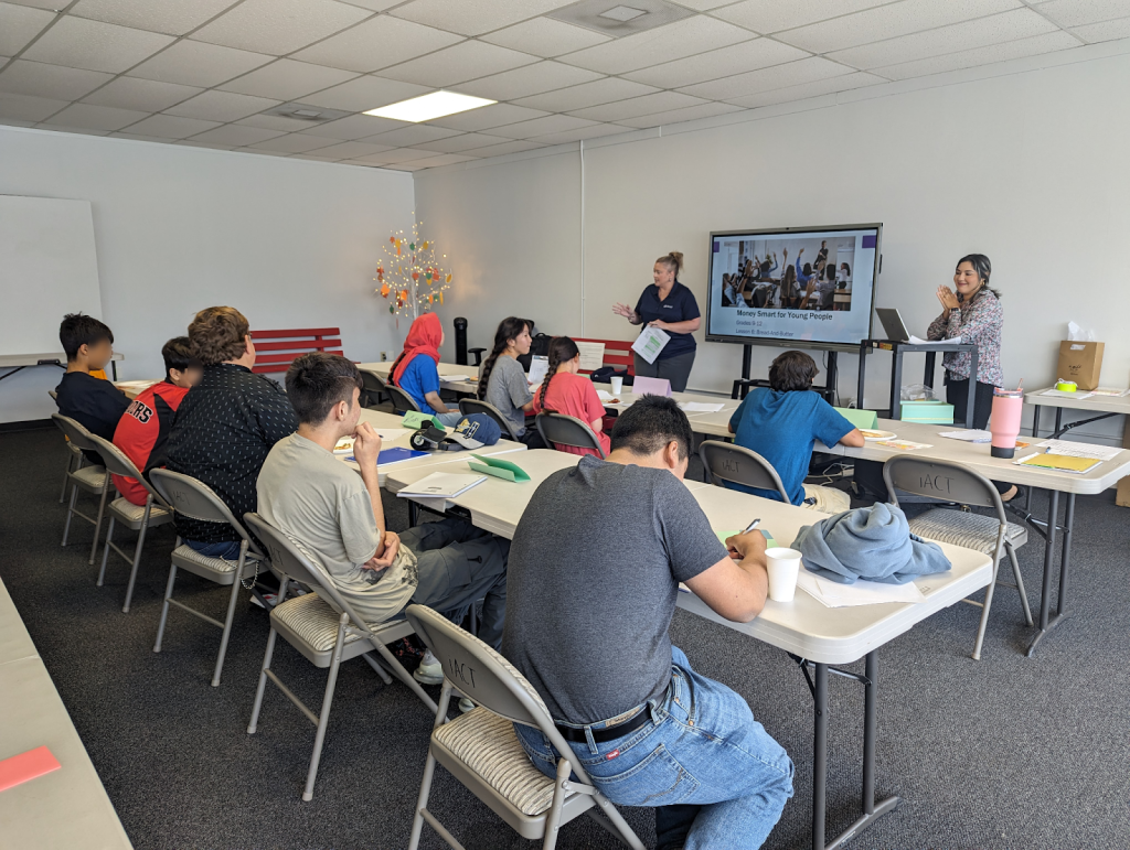 Two presenters stand at the front, pointing to a large screen showing a presentation. Around the table, several students, both young men and women, sit at laptops and notebooks.