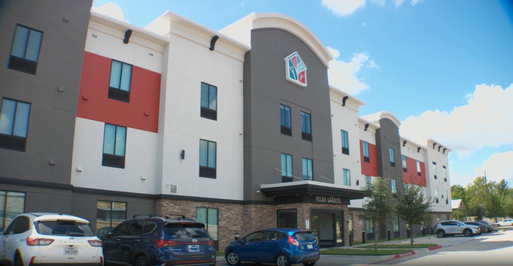 The front view of the Pecan Gardens building, a modern structure with a large logo at the top center, featuring a colorful house and tree design. The building has a gray and white exterior with red accents, along with a few windows and a neatly landscaped area in front.