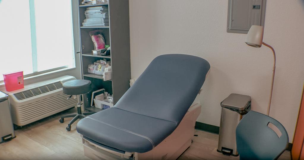 Medical examination room with a blue exam table, shelving with supplies, and a stool next to a window.