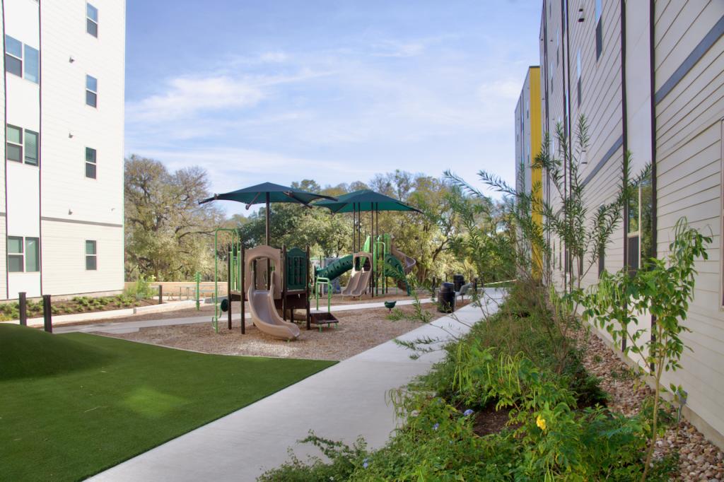 Photos shows a courtyard between apartment buildings with a playground for children surrounded by landscaping.