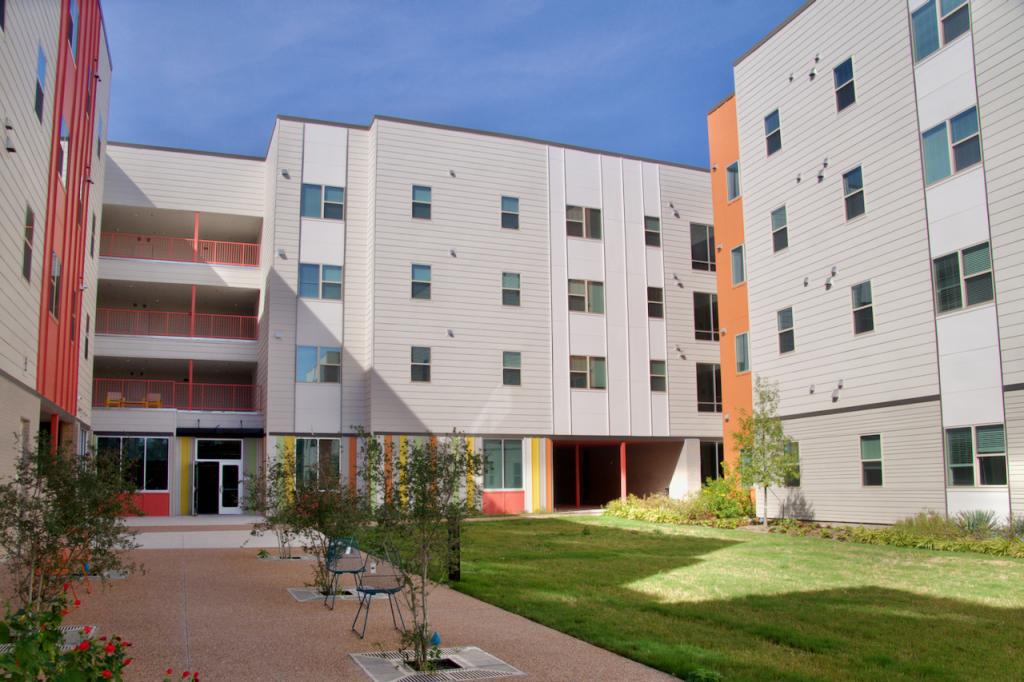 Image shows an inner courtyard surrounded by apartment buildings. Part of the courtyard is covered with grass and features a garden; another part of the courtyard has a playground, a seating area with trees and a playground and picnic area.