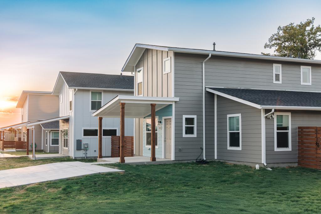 Image shows three two-story homes facing toward the left. Each home has a covered front porch and is clad in vinyl plank siding in different colors such as tan and blue, with the windows and doors trimmed in white.  