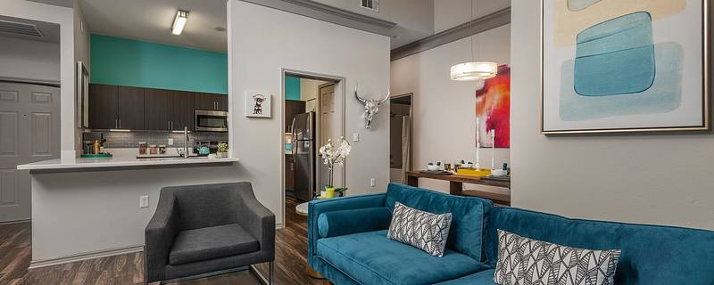 A modern Twelve100 apartment with an interior featuring an open-concept design. The living area includes a teal sofa with patterned throw pillows and a gray armchair on a dark wood floor. The kitchen, visible in the background, has dark cabinets, a white countertop, and stainless-steel appliances.