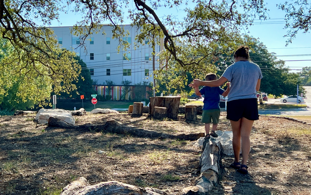 A parent helps a toddler balance walking across a log in a nature play area