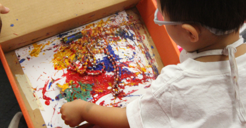 Preschool student experimenting with splatter paint effects and movement using a shoebox shaker.