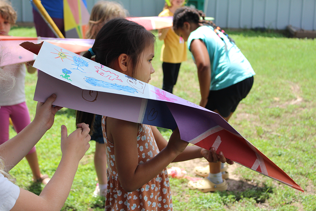 ourdoors on the lawn students prepare to throw giant paper airplanes made of colorful construction paper covered in drawings