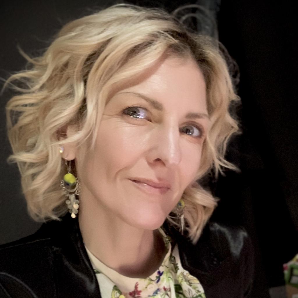 A close up photographic portrait of a person with only their face in view. They are making eye contact with the viewer and smiling.