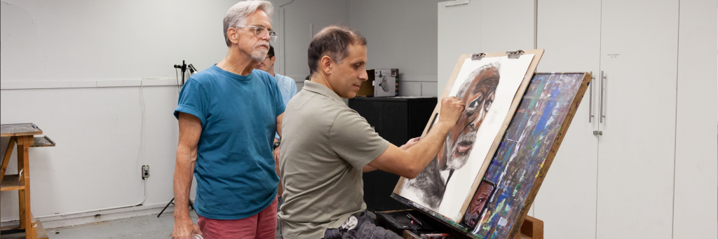 A drawing student working on a drawing while an instructor inspects the artwork.