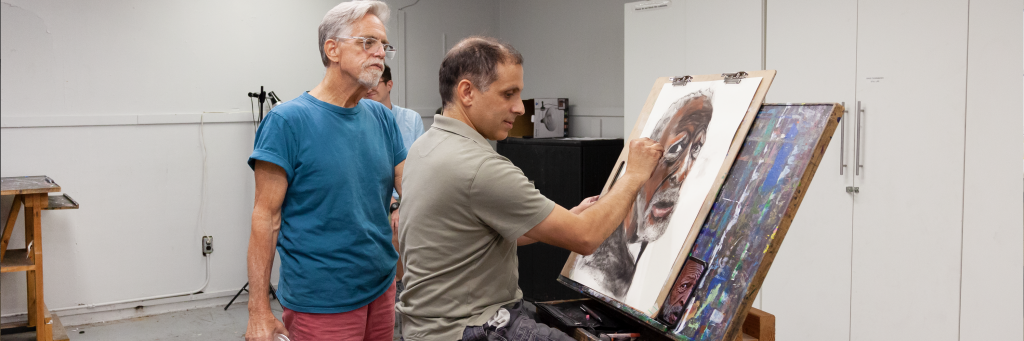A photograph of a student drawing a portrait of a person while an instructor looks on