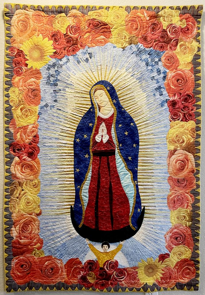 Image of Mary Ann Vaca-Lambert's art quilt "La Virgen de Guadalupe." The Virgin Mary standing, surrounded by golden rays of light. A boarder of red, peach and yellow roses are outlined by a yellow stitched trim.