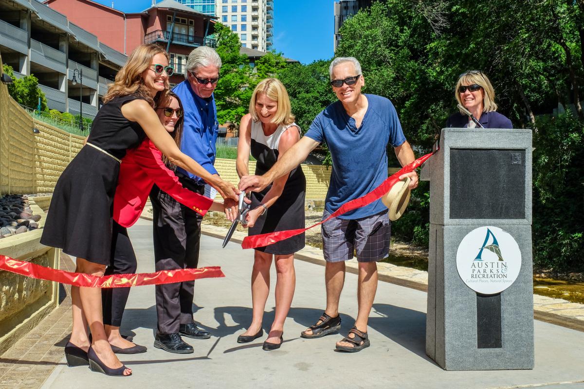 City officials and community leaders cutting a red ribbon near a podium. They are standing on a concrete hike and bike path.