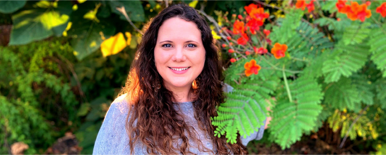 Photograph of Aimee Aubin standing next to trees and red flowers.