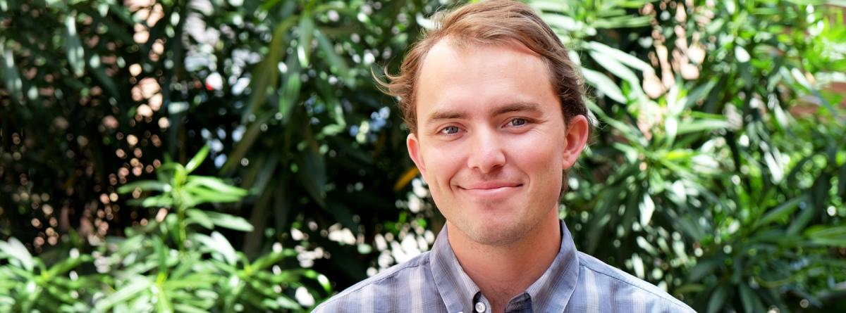 Photo of Brooks Bailey smiling at the camera posed in front of lush green plants. He is wering a purple-ish plaid collared shirt.