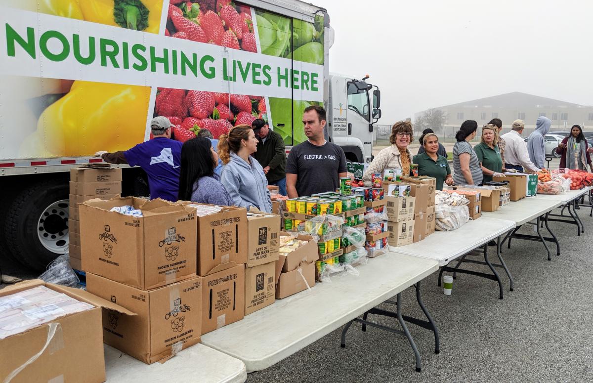 Photo of lots of food on long tables and about 13 volunteers ready to pass out the food. A truck in the background reads "Nourishing Lives Here"