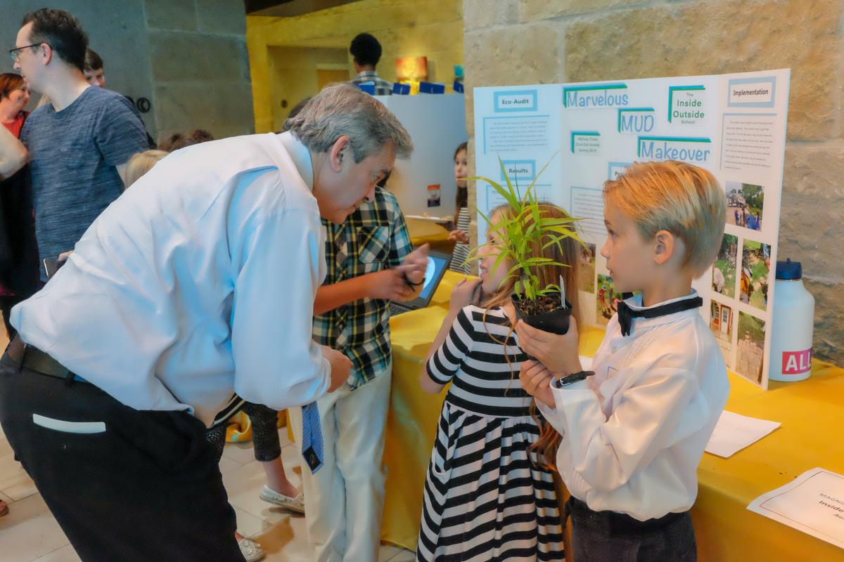 Mayor Steve Adler speaks with students about their sustainability projects. One student is standing nearby holding a green plant.