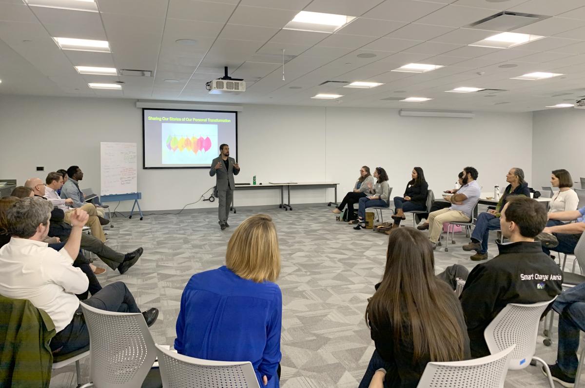 A group of people sitting in a circle in a conference room. They are facing a facilitator and powerpoint screen.