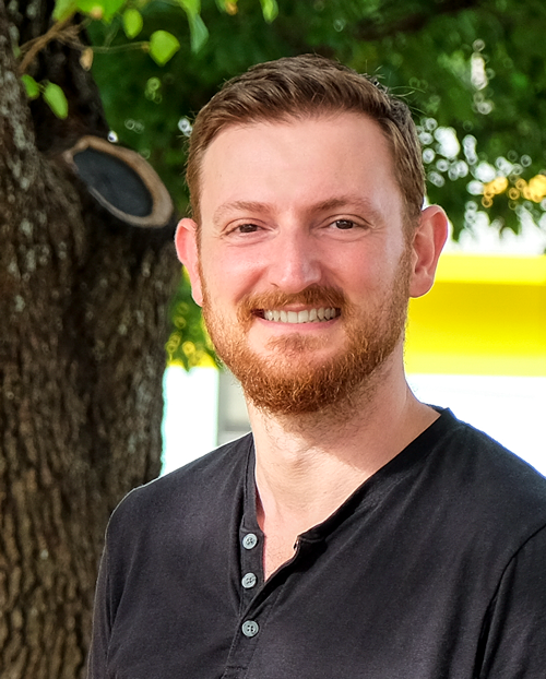 Headshot of Ilya Shmuelenson. He is wearing a black shirt and standing in front of a tree.