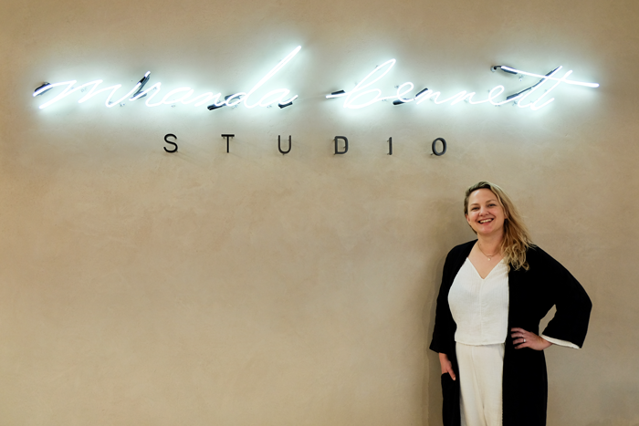 Photo of Miranda below a neon sign that reads "Miranda Bennett Studio" 