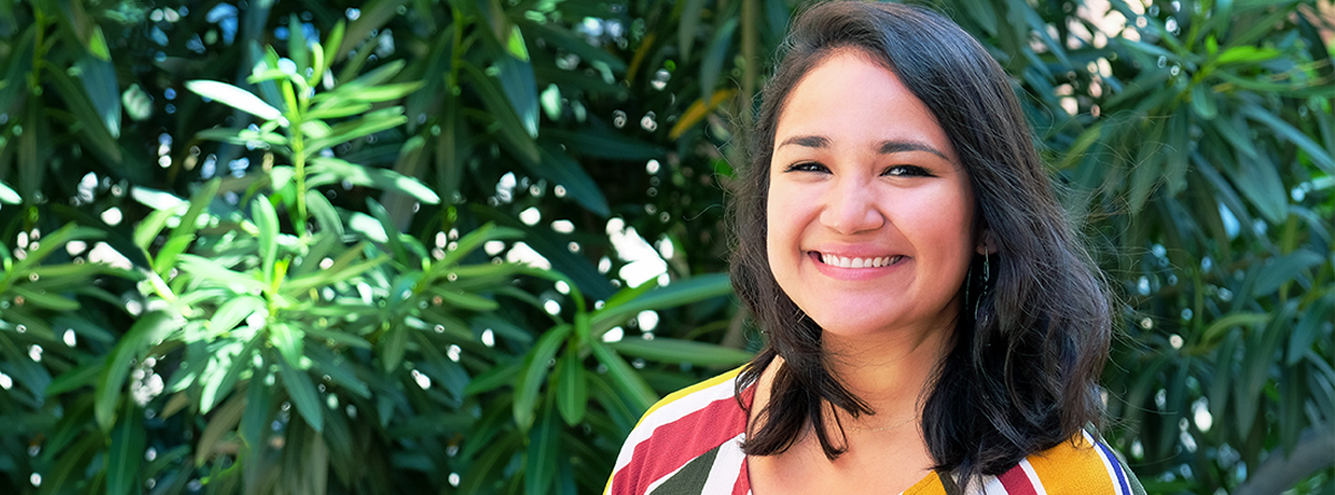 Photo of Phoebe Romero smiling in the background with lush green plants behind her. She is wearing a yellow, red, white, and blue striped top.