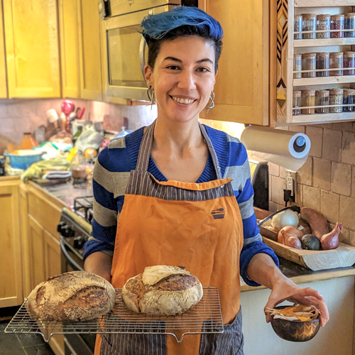 Rosie in an orange apron standing in her kitchen. She is holding a couple loaves of fresh-baked sourdough bread and a cup of candied orange peels.