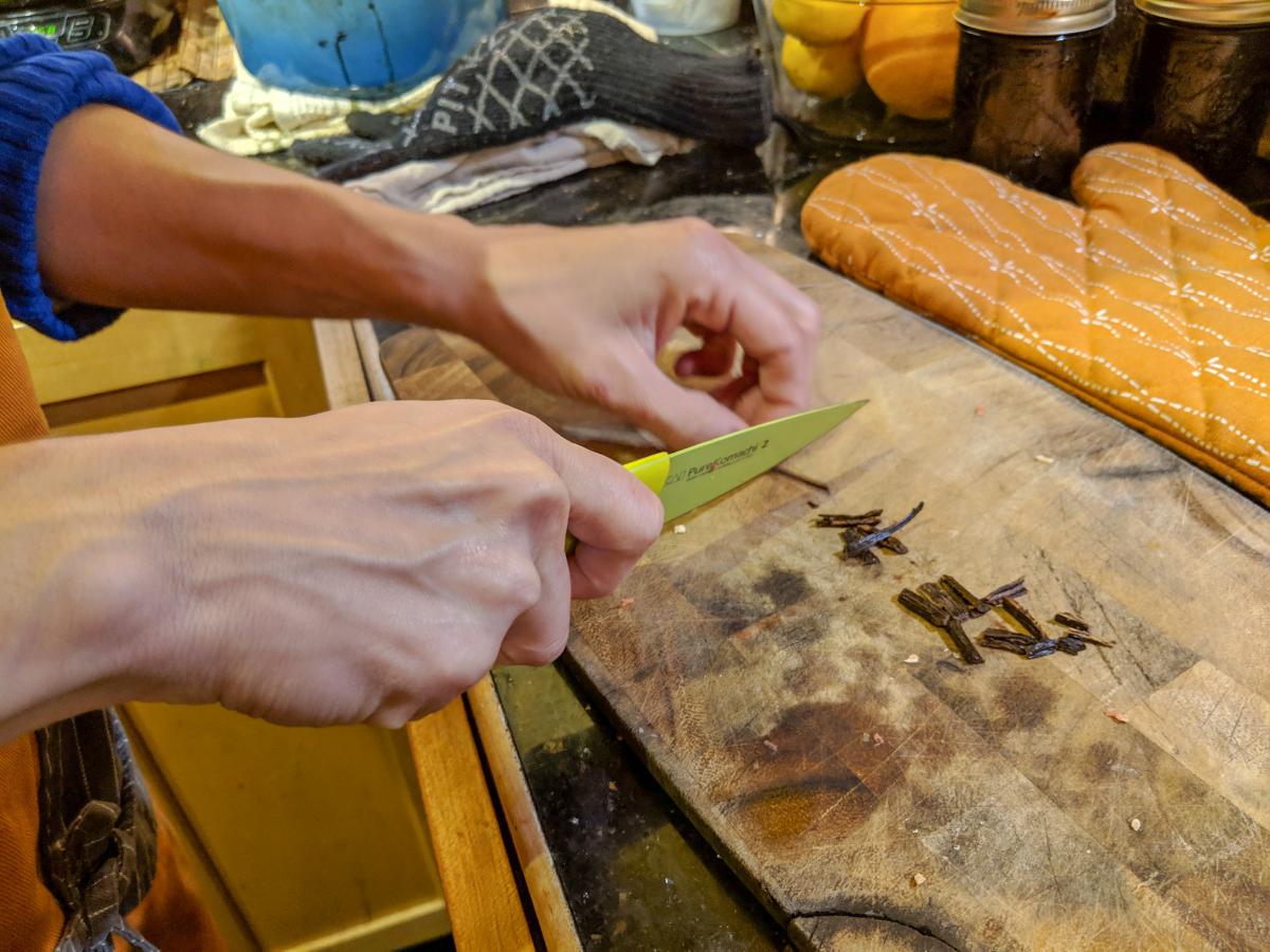 Closeup of hands and a cutting board cutting vanilla pods into small pieces with a bright yellow knife.