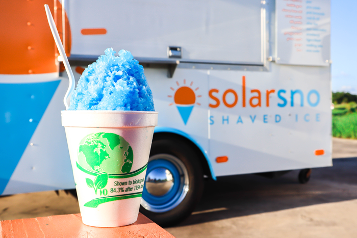 Sno-cone with blue shaved ice and spoon in foreground. Cup reads "shown to be biodegradable" and has a green picture of earth on it. The Solar Sno shaved ice trailer is in the background.