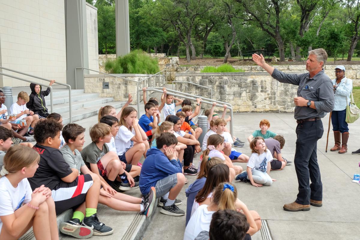 Teacher Greg Mele explaining a project to students. Students are all seated in front of a building on some steps.