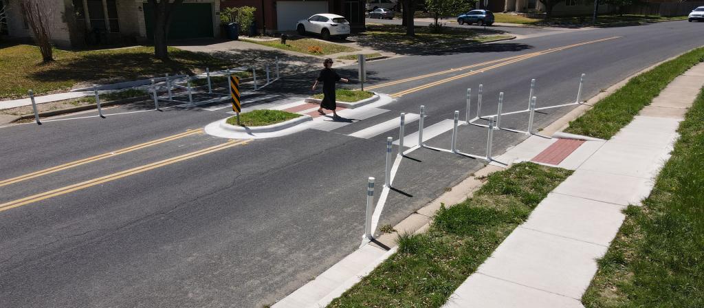 A pedestrian crosses Palace Parkway using a marked crosswalk featuring a crossing island and flex posts to encourage reduced vehicular speeds through the area.