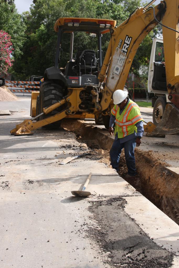 A worker stands inside an open trench on the ground. Behind him a yellow excavator is stationary.