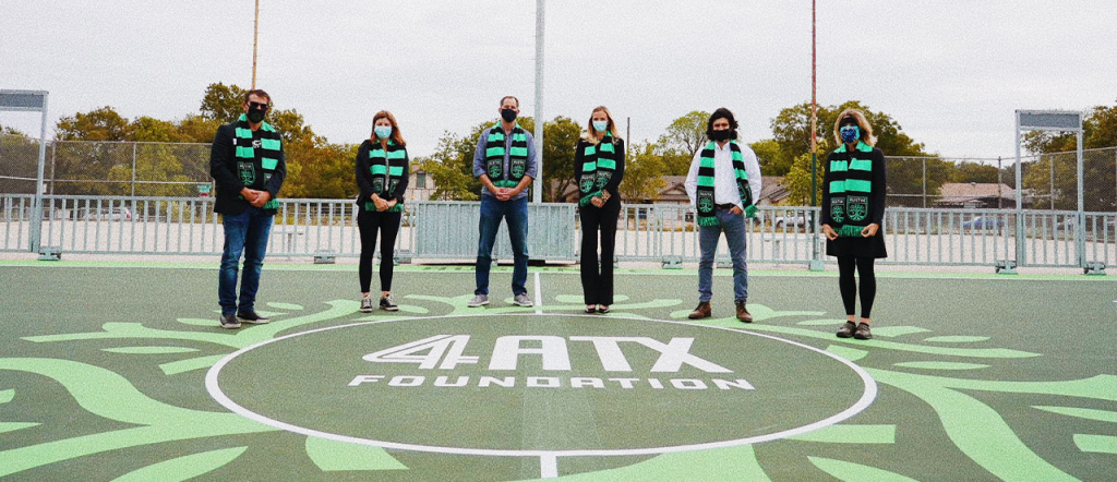 6 people stand in a semi-circle on a green pitch. The pitch has words reading "4 ATX Foundation"