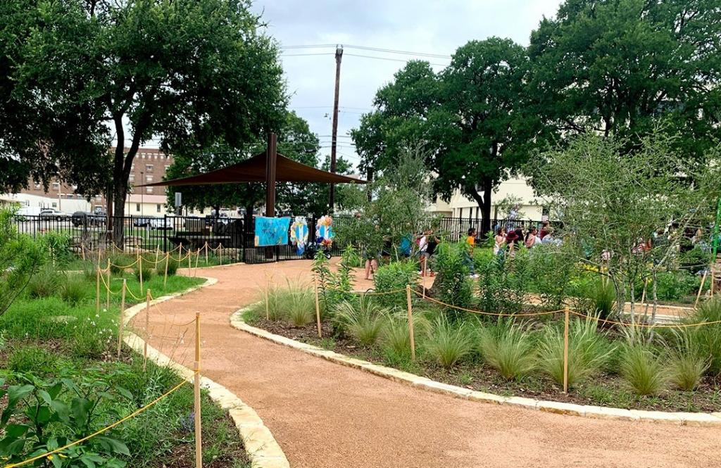 A gravel path winds between two sections of the garden. The garden sections have grass, short trees, and flowers. A group of people can be seen through the greenery in the background of the photo.