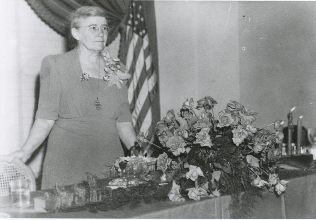 Blanton standing behind table with flowers. American flag behind her. 