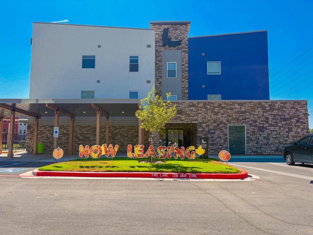 Rendering shows several multi-story buildings that range in color from grey to orange to blue to purple, surrounded by a parking lot, trees and grass.