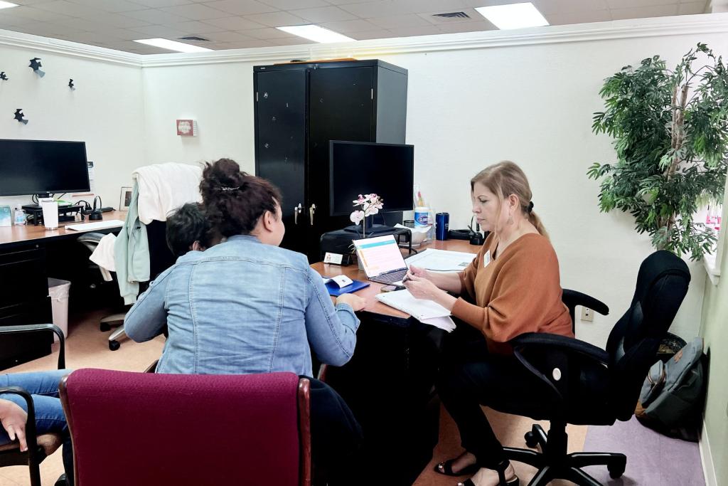Image shows a woman sitting at a desk, helping a family fill out paperwork. 