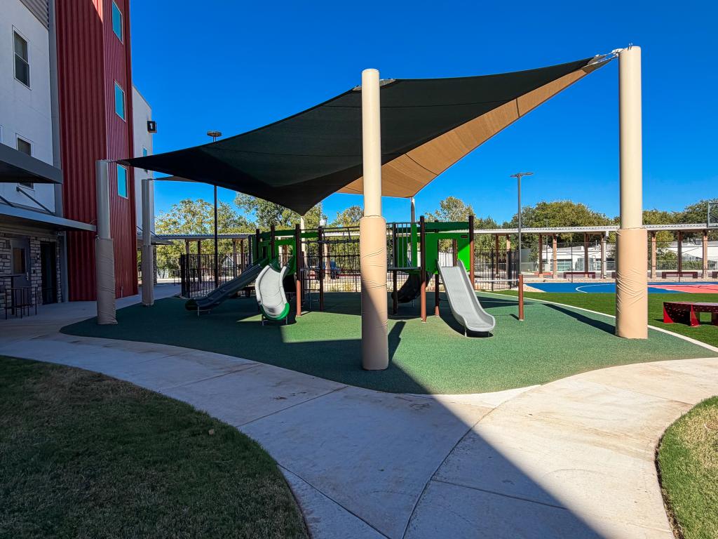 View of the playground showing slides and climbing equipment under a tan and green shade canopy, with nearby walkways and a basketball court in the background.