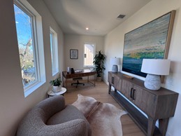 Small home office with three tall windows, a wooden desk and chair, a cushioned armchair, indoor plants, and a sideboard with two lamps beneath a large landscape painting.
