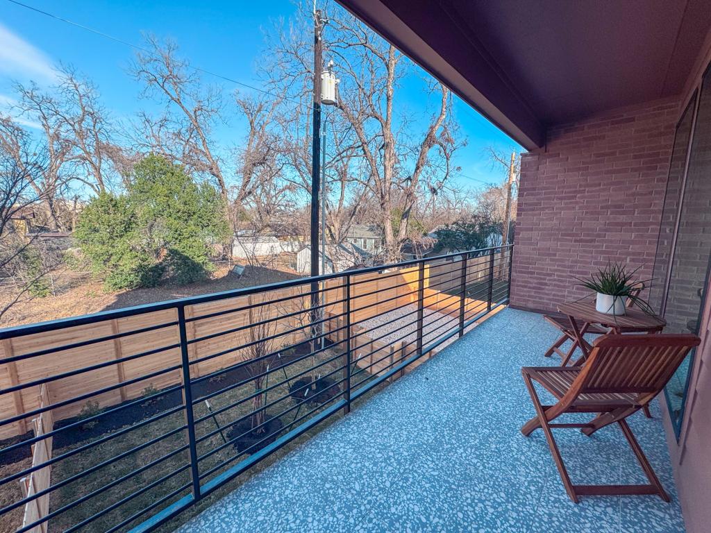 Private balcony with terrazzo-style flooring, modern metal railing, and seating, overlooking trees and a fenced yard on a clear day.