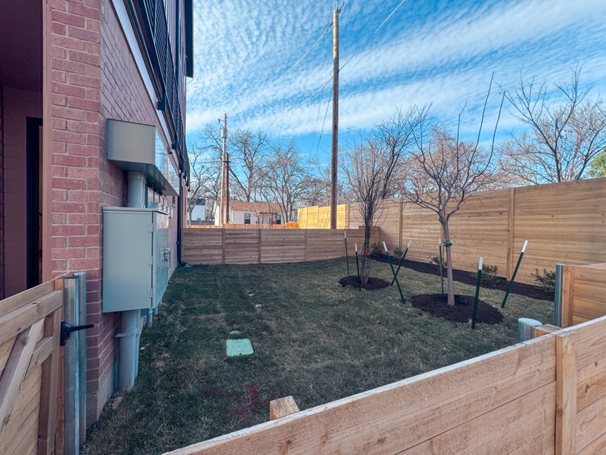 Fenced backyard next to a two-story brick building with a small grass lawn, two young trees supported by stakes, and wooden privacy fencing on all sides.