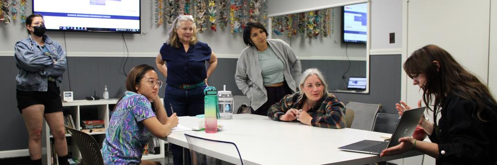 A room with six people around a table. One person is giving a presentation on their laptop while the other five people are observing.