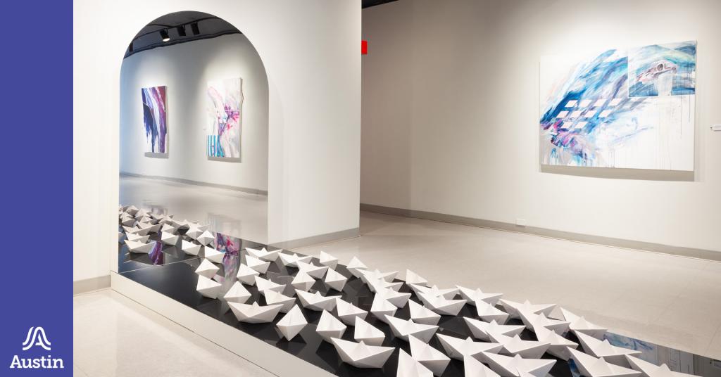 A photograph of a gallery space with an art installation of boats on the floor and paintings on the wall and a graphic element on the left of the image of the Austin A logo.