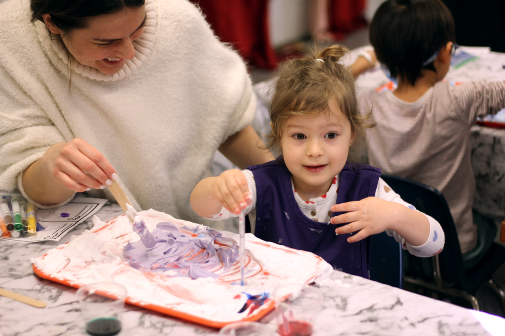 Photo of smiling caregiver and child smiling and working together swirling colors on a tray of shaving cream 
