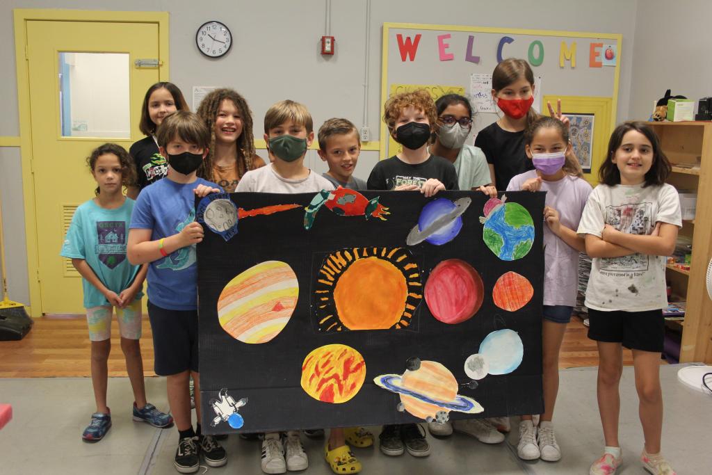Group of students smiling and posing with their solar system group project with painted paper sun and planets on a larger sheet of black paper.