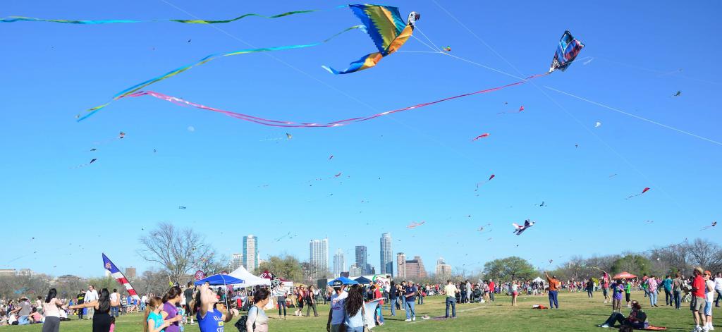 "image of adults and youth flying kites in a large park with Austin downtown skyline in the background"