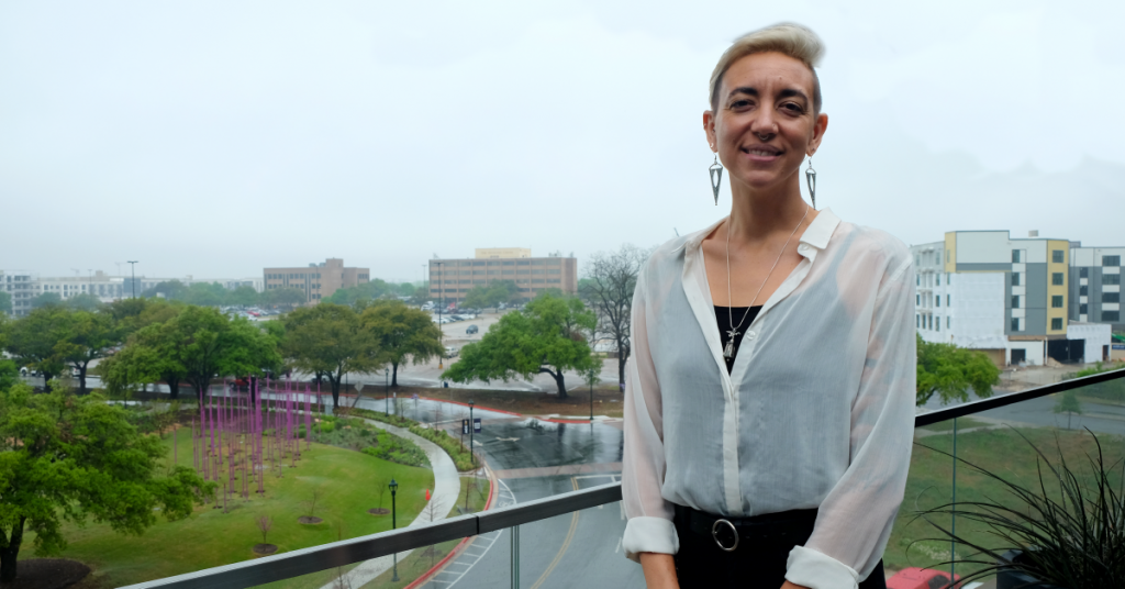 Angela Baucom smiles on a balcony with a view of a park and buildings behind her.