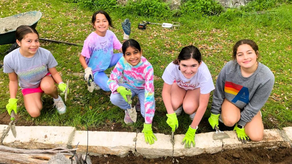 Five students sit in a row in front of a plant bed with garden tools.