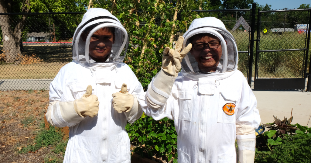 Two young boys wear beekeeper suits and smile for the camera.