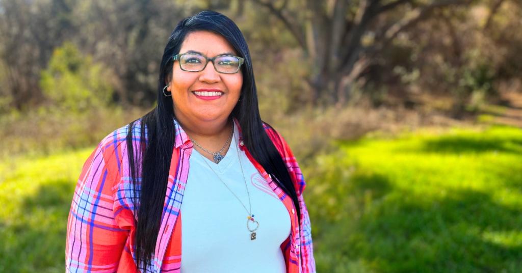 Leti Alvarez stands outside smiling. She wears a red flannel and stands in front of a field with trees.